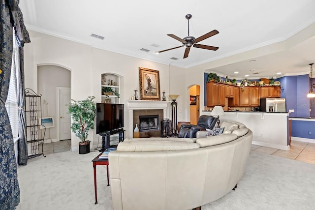 living room featuring a tiled fireplace, ceiling fan, light tile patterned floors, ornamental molding, and built in features