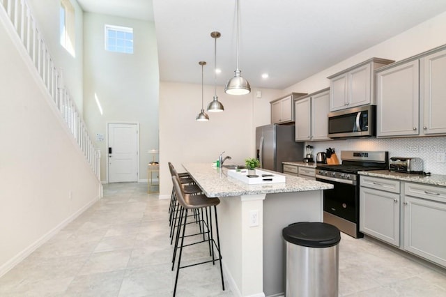 kitchen featuring appliances with stainless steel finishes, a center island with sink, light stone counters, hanging light fixtures, and light tile patterned flooring