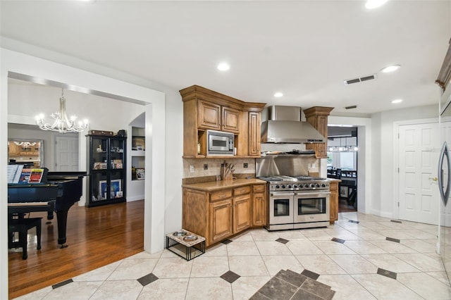 kitchen featuring light tile patterned flooring, appliances with stainless steel finishes, wall chimney range hood, and a notable chandelier