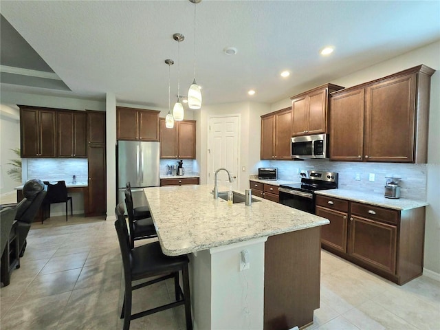 kitchen with sink, a kitchen island with sink, stainless steel appliances, decorative backsplash, and decorative light fixtures