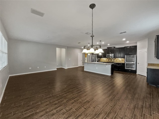 kitchen featuring hanging light fixtures, appliances with stainless steel finishes, dark hardwood / wood-style flooring, a kitchen island with sink, and backsplash