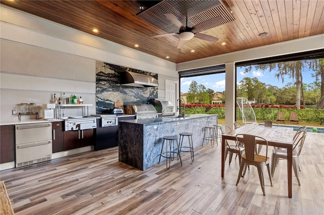 kitchen featuring wood ceiling, wall chimney range hood, an island with sink, sink, and light wood-type flooring