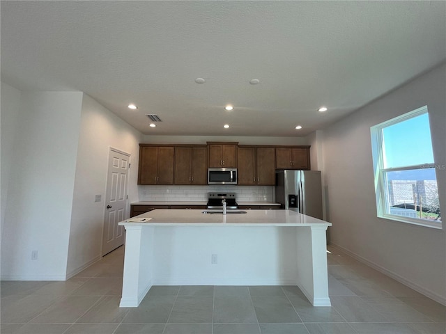 kitchen with an island with sink, sink, appliances with stainless steel finishes, light tile patterned floors, and backsplash