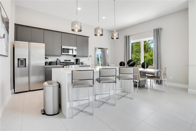 kitchen with light tile patterned flooring, gray cabinets, stainless steel appliances, hanging light fixtures, and a center island with sink