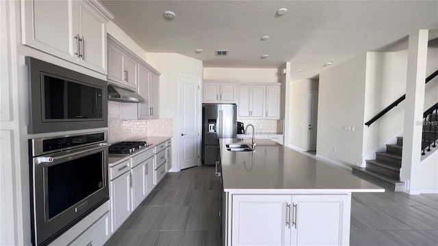 kitchen featuring tasteful backsplash, a kitchen island with sink, under cabinet range hood, stainless steel appliances, and a sink