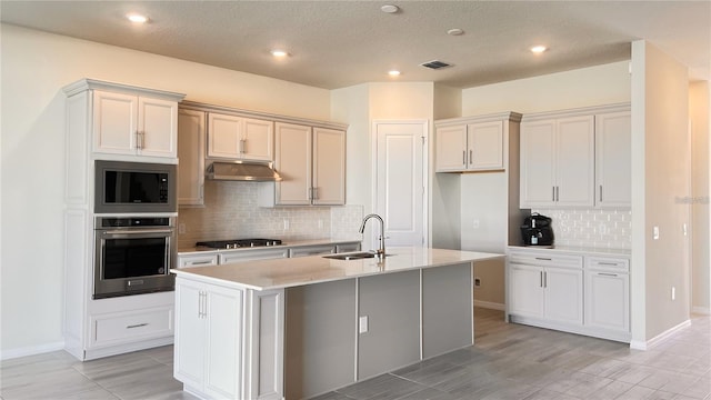 kitchen featuring an island with sink, stainless steel appliances, sink, backsplash, and a textured ceiling