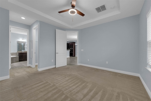 unfurnished bedroom with a raised ceiling, ensuite bathroom, light colored carpet, and ceiling fan