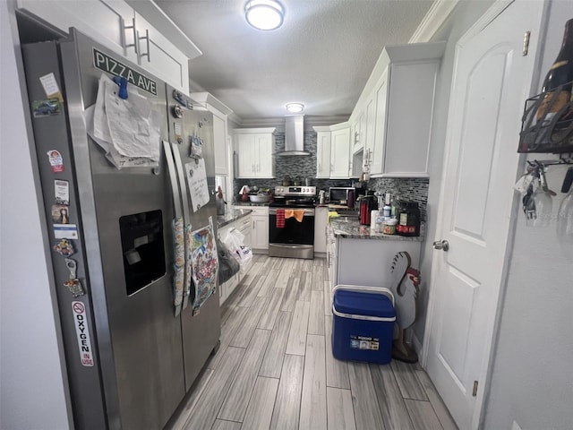 kitchen with wall chimney range hood, dark stone countertops, white cabinets, stainless steel appliances, and backsplash