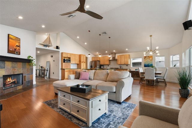 living room with high vaulted ceiling, dark hardwood / wood-style flooring, a tiled fireplace, and ceiling fan with notable chandelier