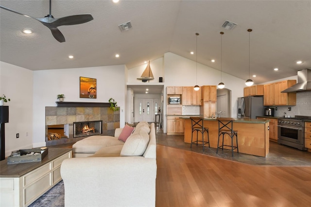 kitchen with wall chimney range hood, dark hardwood / wood-style flooring, ceiling fan, stainless steel appliances, and a tiled fireplace