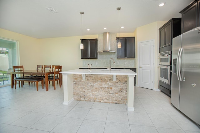 kitchen featuring wall chimney exhaust hood, dark brown cabinets, hanging light fixtures, stainless steel appliances, and a kitchen island with sink