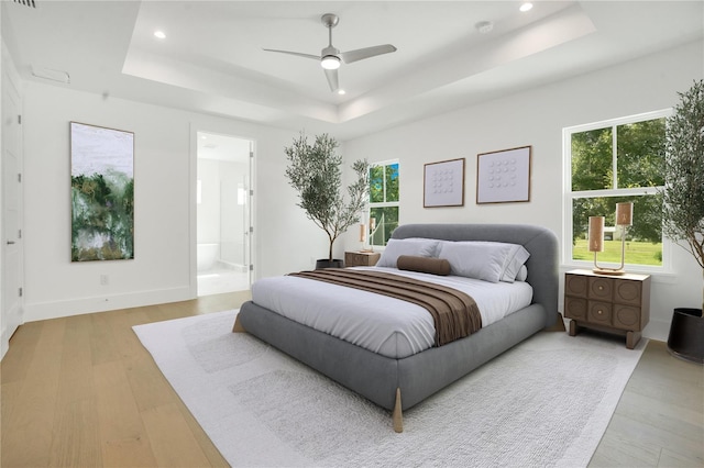 bedroom featuring ceiling fan, ensuite bathroom, a raised ceiling, and light wood-type flooring