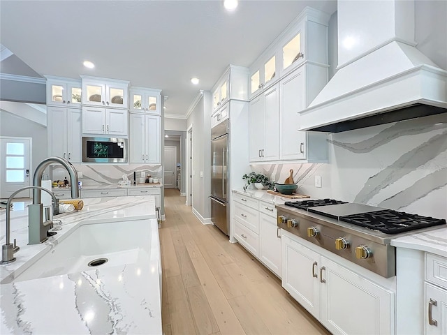 kitchen with custom exhaust hood, white cabinets, light stone countertops, built in appliances, and sink