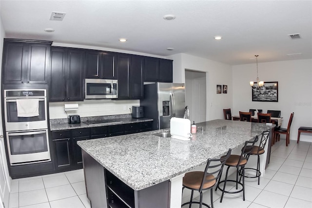 kitchen with light tile patterned flooring, an island with sink, pendant lighting, stainless steel appliances, and light stone countertops