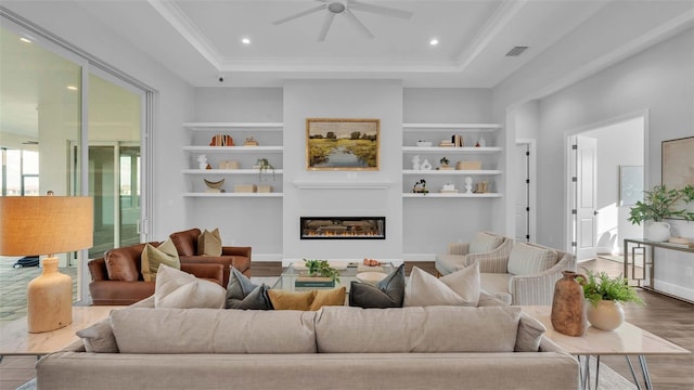 living room with ceiling fan, a raised ceiling, built in features, hardwood / wood-style flooring, and crown molding