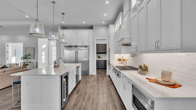 kitchen with built in appliances, an island with sink, beverage cooler, white cabinetry, and hanging light fixtures