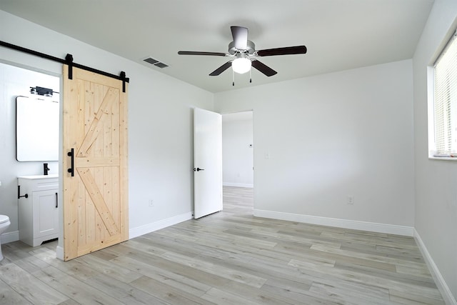 unfurnished bedroom with light hardwood / wood-style floors, ensuite bath, a barn door, and ceiling fan