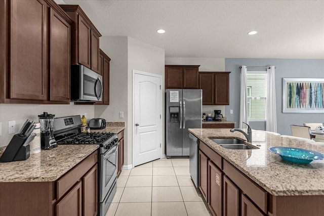 kitchen featuring sink, light tile patterned floors, appliances with stainless steel finishes, light stone countertops, and a center island with sink