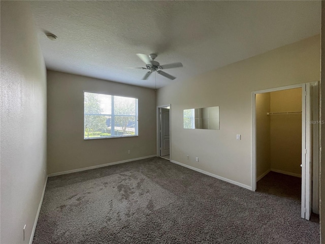 unfurnished bedroom with ceiling fan, a walk in closet, a textured ceiling, and dark colored carpet