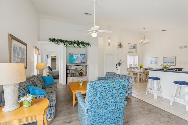 living area with visible vents, a towering ceiling, light wood finished floors, and ceiling fan with notable chandelier