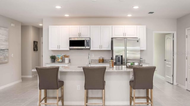 kitchen with light tile patterned flooring, white cabinetry, light stone countertops, appliances with stainless steel finishes, and a kitchen island with sink