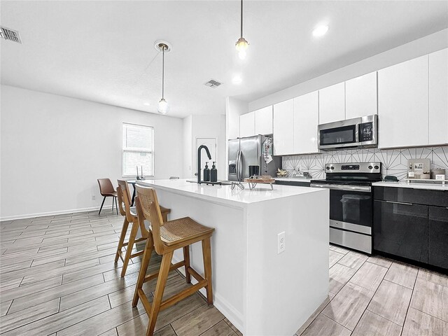 kitchen featuring appliances with stainless steel finishes, an island with sink, white cabinets, and decorative light fixtures