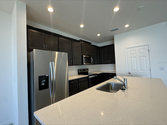 kitchen with sink, light stone counters, an island with sink, a textured ceiling, and appliances with stainless steel finishes