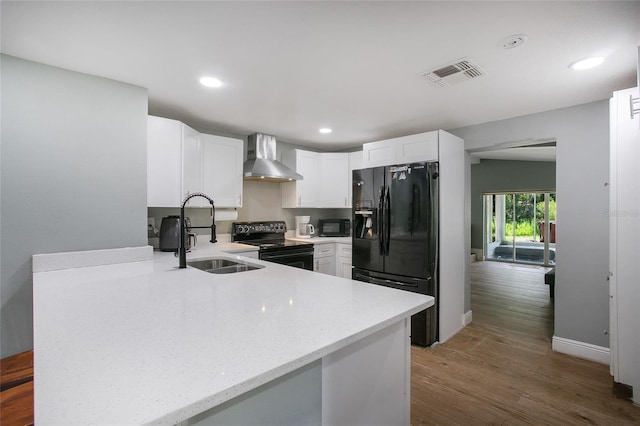 kitchen with white cabinets, a sink, black appliances, a peninsula, and wall chimney exhaust hood