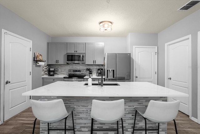 kitchen with gray cabinetry, light stone countertops, a kitchen breakfast bar, an island with sink, and appliances with stainless steel finishes