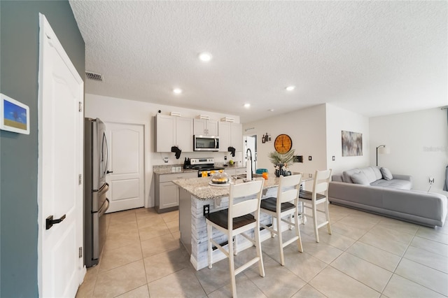 kitchen featuring a center island with sink, a textured ceiling, light tile patterned flooring, light stone counters, and stainless steel appliances