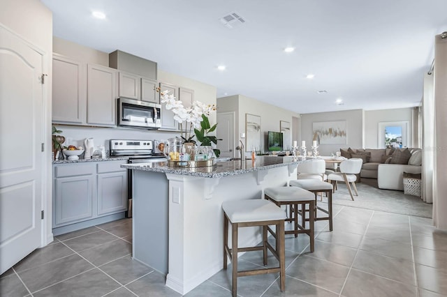 kitchen featuring tile patterned floors, stainless steel appliances, a breakfast bar area, a center island with sink, and gray cabinetry