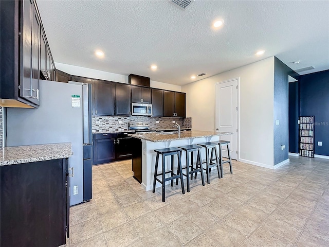 kitchen featuring a kitchen island with sink, stainless steel appliances, light stone counters, a breakfast bar area, and decorative backsplash