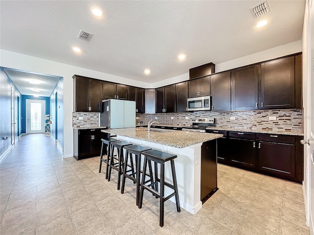 kitchen with a kitchen bar, appliances with stainless steel finishes, light stone counters, dark brown cabinetry, and a kitchen island with sink