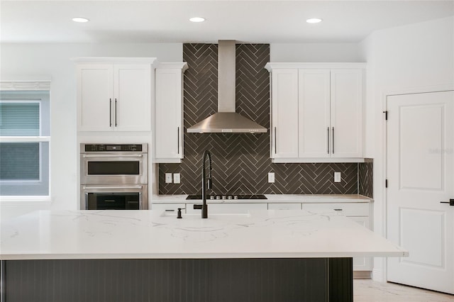 kitchen with wall chimney range hood, white cabinets, a kitchen island with sink, and double oven