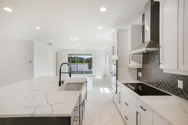 kitchen featuring black electric stovetop, wall chimney range hood, sink, an island with sink, and white cabinetry