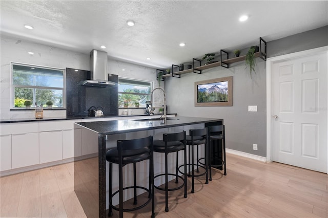 kitchen featuring a breakfast bar area, white cabinetry, wall chimney range hood, light hardwood / wood-style flooring, and a center island with sink