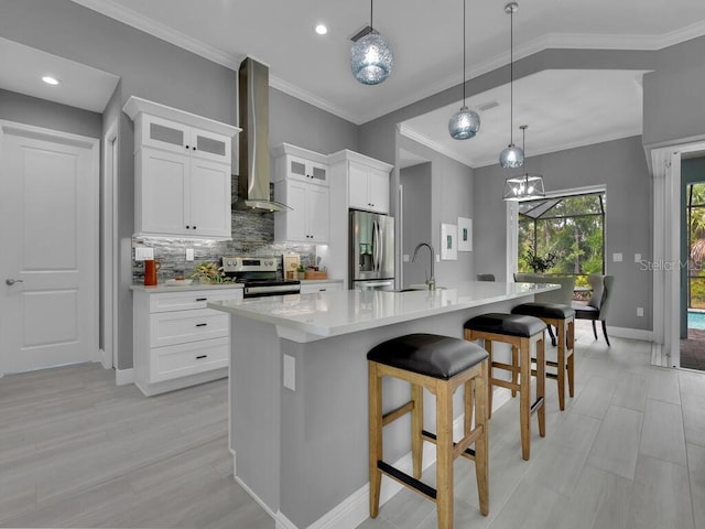 kitchen with an island with sink, decorative light fixtures, white cabinets, appliances with stainless steel finishes, and wall chimney range hood