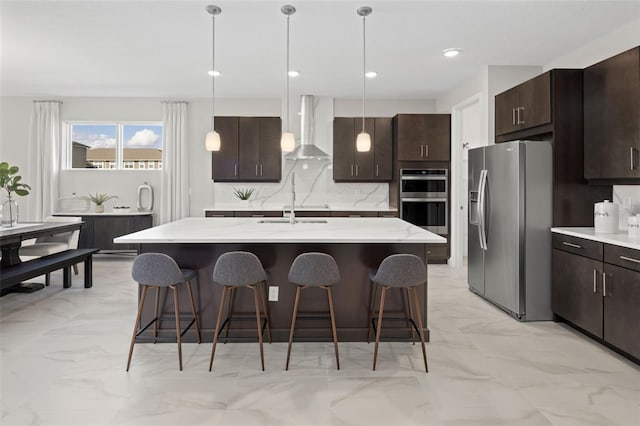 kitchen with wall chimney exhaust hood, sink, decorative light fixtures, stainless steel appliances, and a kitchen island with sink