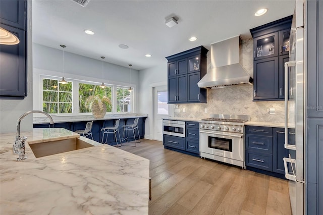 kitchen with premium appliances, light stone counters, glass insert cabinets, a sink, and wall chimney range hood