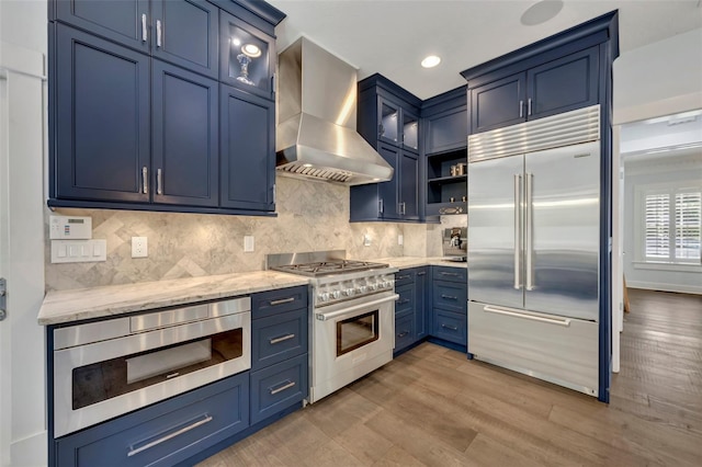 kitchen featuring built in appliances, light stone counters, wall chimney range hood, blue cabinetry, and glass insert cabinets