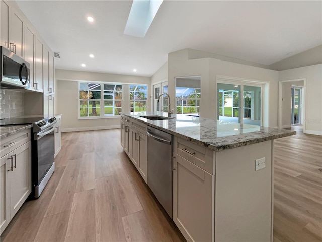 kitchen with lofted ceiling with skylight, an island with sink, sink, stainless steel appliances, and light stone countertops