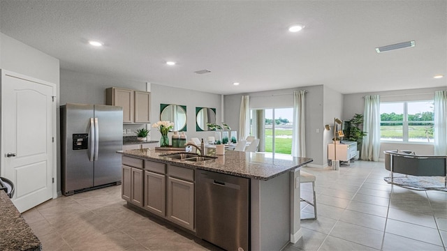 kitchen with appliances with stainless steel finishes, dark stone counters, sink, light tile patterned floors, and a kitchen island with sink