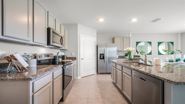 kitchen with light tile patterned floors, gray cabinets, sink, appliances with stainless steel finishes, and a center island with sink