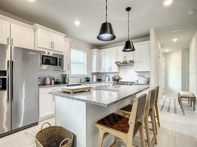 kitchen with light stone counters, under cabinet range hood, stainless steel appliances, white cabinets, and decorative light fixtures