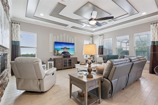 living room with a raised ceiling, a wealth of natural light, ceiling fan, and a stone fireplace