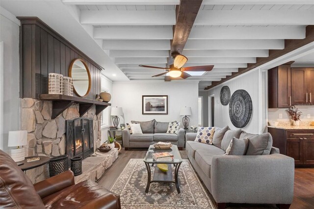 living room with beam ceiling, a stone fireplace, dark wood-type flooring, and ceiling fan