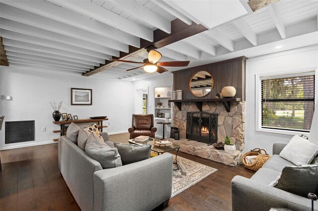 living room with ceiling fan, dark wood-type flooring, a stone fireplace, and beam ceiling