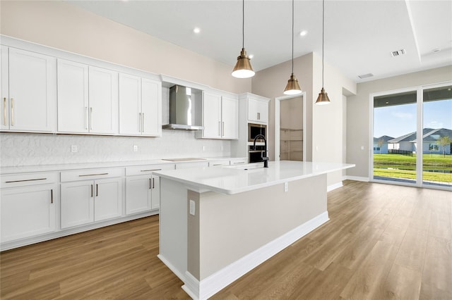 kitchen featuring hanging light fixtures, white cabinetry, wall chimney exhaust hood, light hardwood / wood-style flooring, and a kitchen island with sink