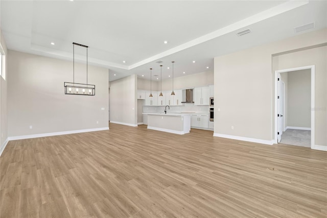 unfurnished living room featuring an inviting chandelier, sink, light wood-type flooring, and a raised ceiling