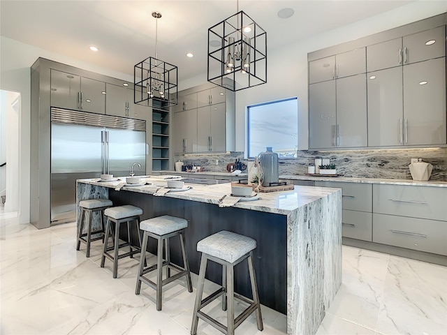 kitchen with gray cabinets, an island with sink, built in refrigerator, light stone countertops, and decorative light fixtures